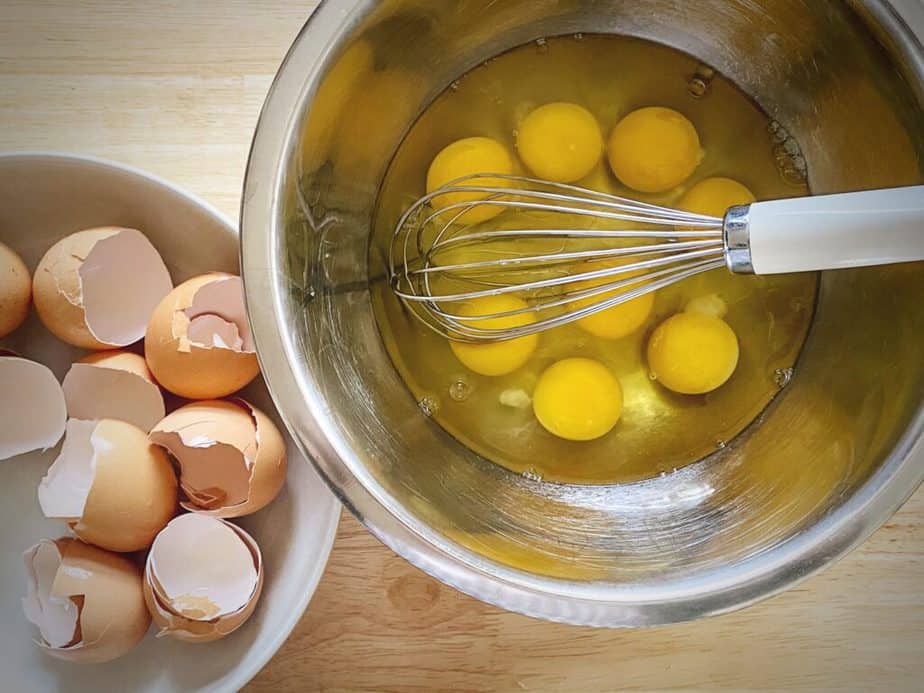 eggs in a mixing bowl with a whisk and a short bowl to the side holding egg shells