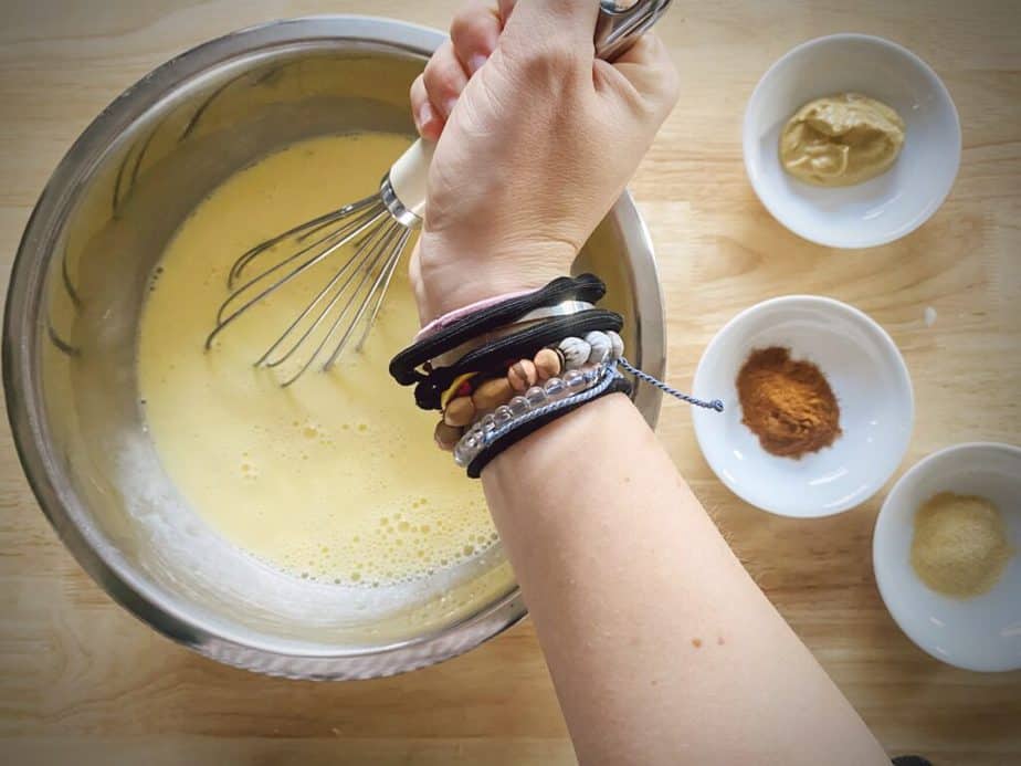 hand whisking custard mixture in silver mixing bowl with cayenne, dijon and garlic powder in small bowls to the side