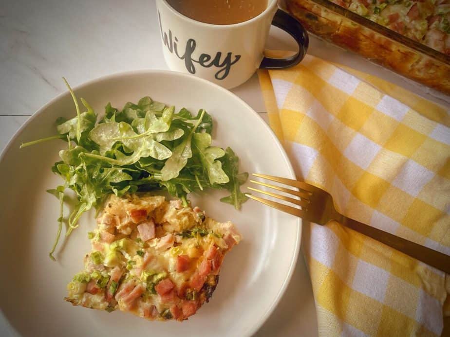 overhead shot of plated strata with side salad on a white plate with a gold fork, yellow and white gingham napkin, mug of coffee and remaining breakfast casserole in 9 x 13 in the background