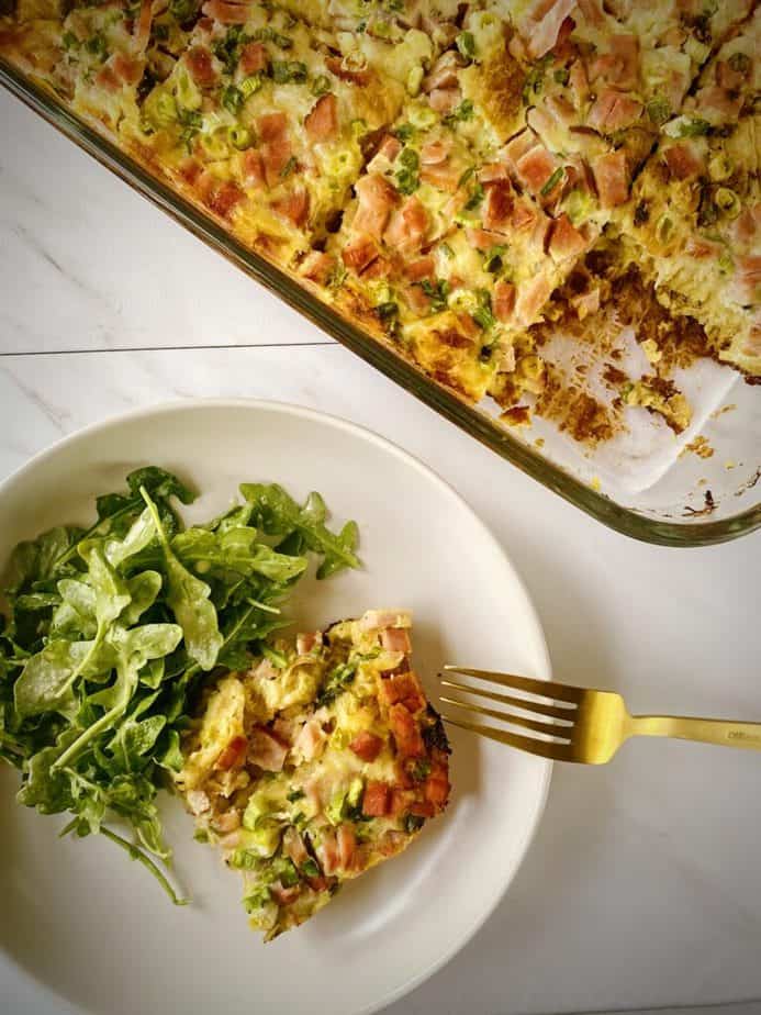 overhead shot of a plated slice of ham and swiss strata with a side salad on a white plate with a gold fork and the remaining breakfast casserole in a 9 x 13