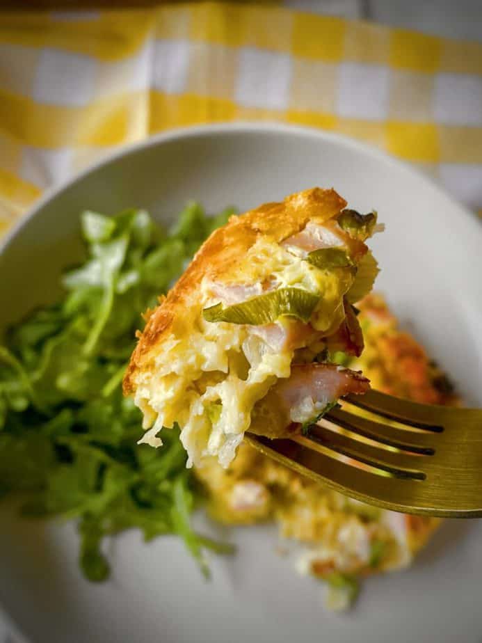 single bite of ham and swiss strata on a gold fork above a plate of strata with side salad atop a yellow and white gingham napkin