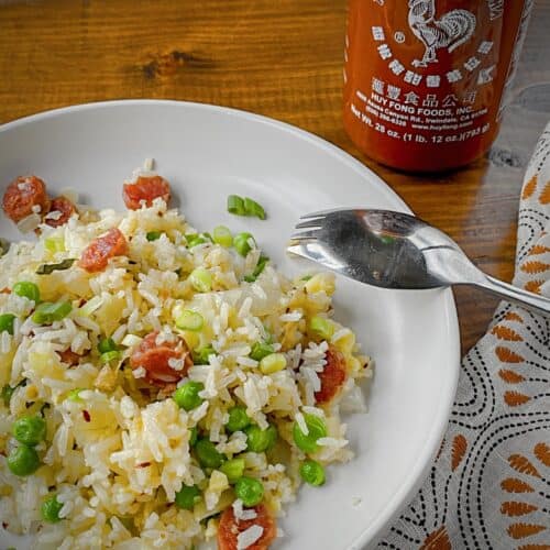 breakfast fried rice with chinese sausage plated in a white pasta bowl with a bottle of sriracha in the back ground and a silver spork with a orange, black and white patterned napkin