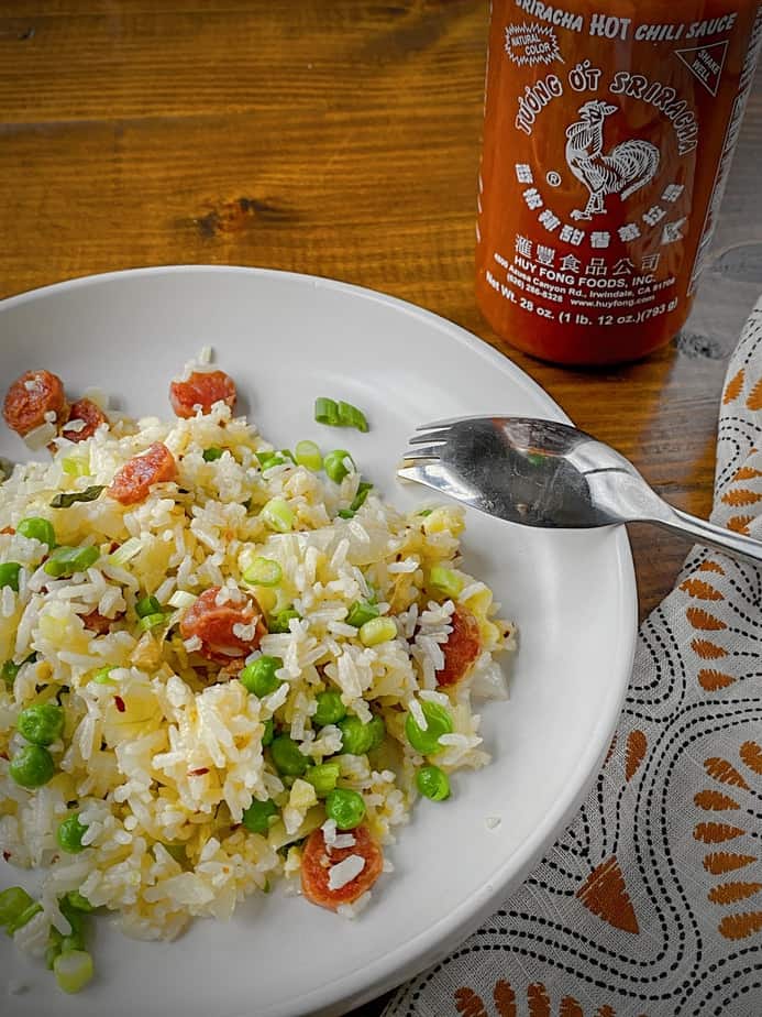breakfast fried rice with chinese sausage plated in a white pasta bowl with a bottle of sriracha in the back ground and a silver spork with a orange, black and white patterned napkin