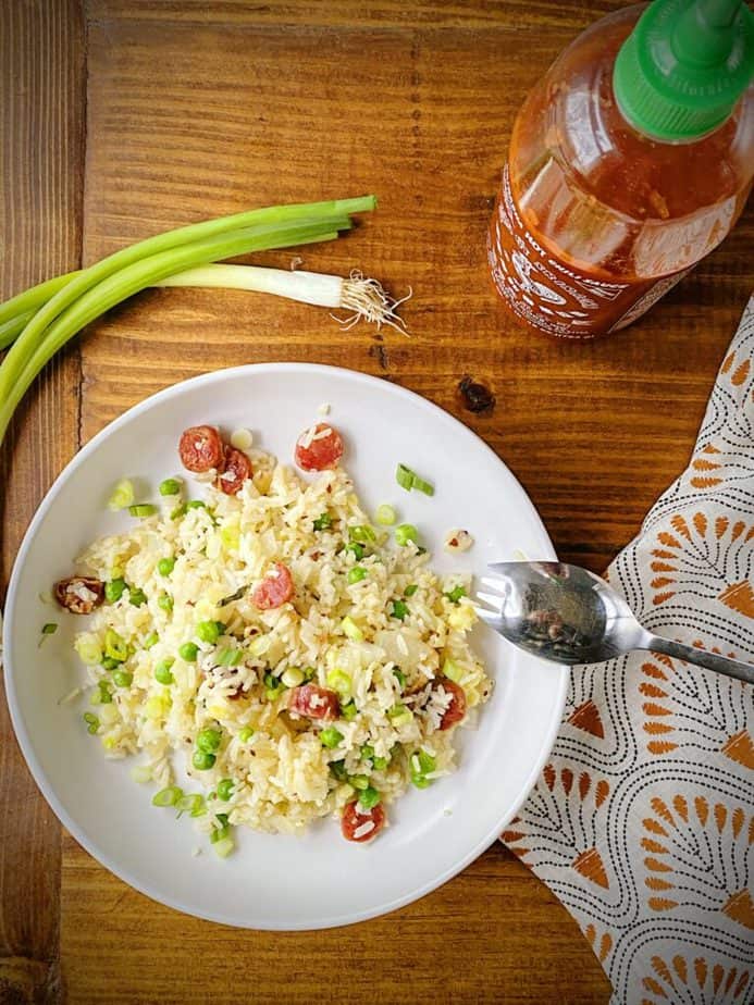 overhead shot of breakfast fried rice, two whole scallions, a bottle of sriracha and a silver spork on a wooden table