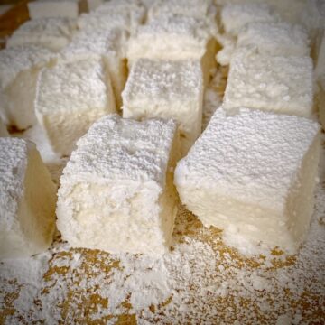 Marshmallows tossed in powdered sugar and corn starch on a wooden cutting board after being cut by a bench scraper.