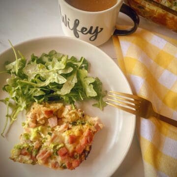 ham and swiss strata slice on a white plate with a green salad, gold fork, cup of coffee and breakfast casserole dish in the background.