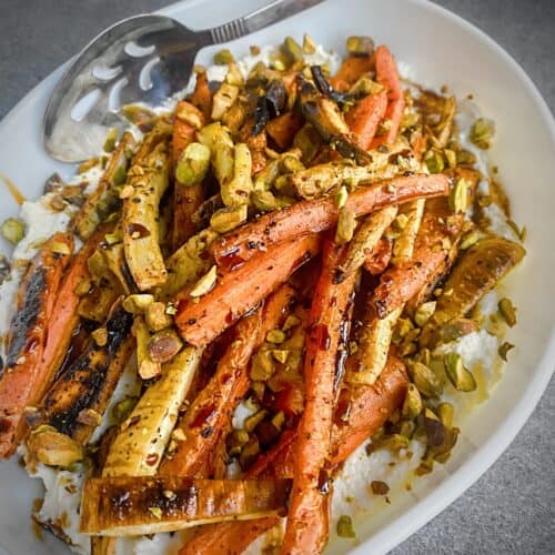 sumac roasted carrots and parsnips on a bed of whipped feta topped with chopped pistachios and a drizzle of pomegranate molasses in a white oval plate