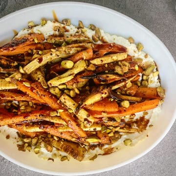sumac roasted carrots and parsnips on a bed of whipped feta topped with chopped pistachios and a drizzle of pomegranate molasses in a white oval plate.