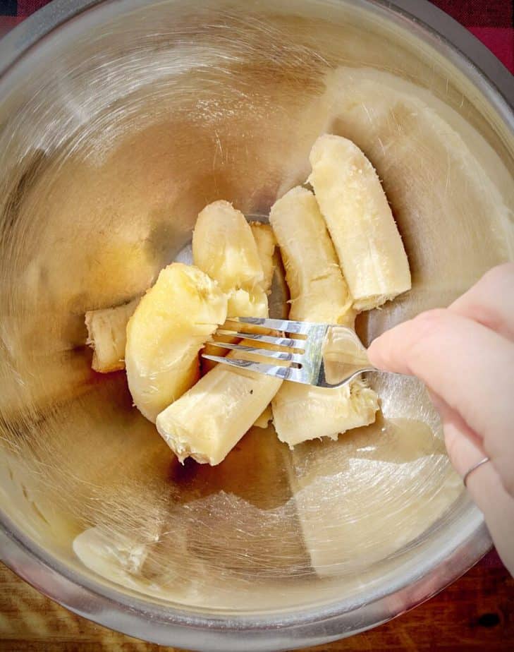 whole bananas in mixing bowl with a hand holding a fork beginning to mash them