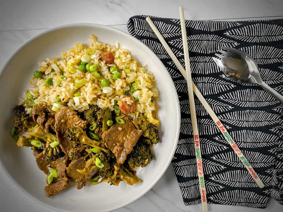 overhead shot of plated broccoli beef in a white pasta bowl with a black and white napkin, chopsticks and a silver spork