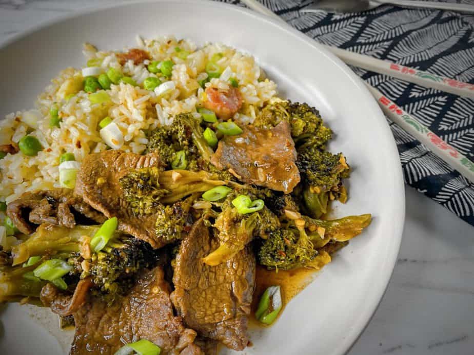 45 degree angle shot of plated broccoli beef with fried rice in a white pasta bowl with a pair of chopsticks and a silver spork on a black and white napkin