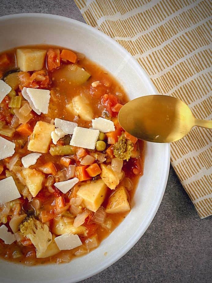 overhead hero shot of instant pot vegetable soup garnished with cheese in a white pasta bowl with a gold spoon on a grey table with a white and mustard yellow napkin