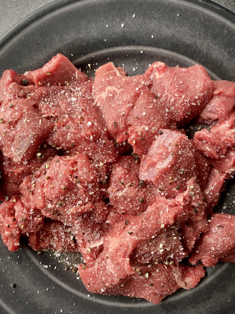raw beef stew meat on a grey plate after being seasoned with salt and pepper