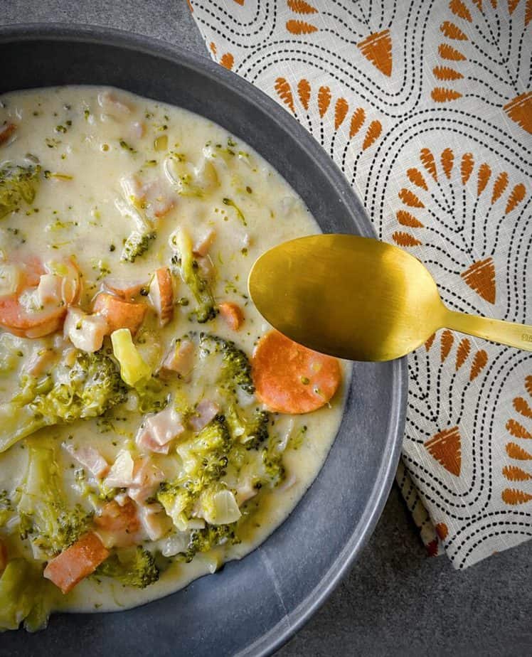 overhead shot of broccoli cheese soup with ham in a grey pasta bowl with a gold spoon and a patterned orange, black and white linen napkin