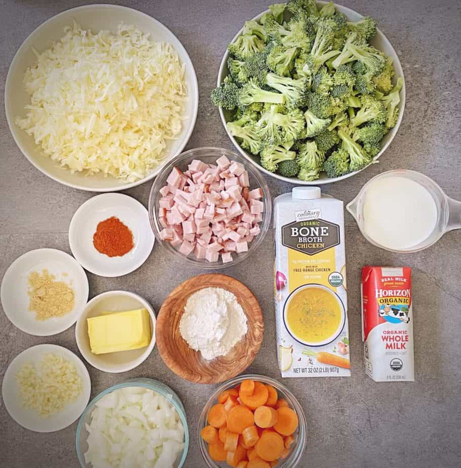 mise en place for broccoli cheese soup recipe - shredded cheddar, broccoli florets, cubed ham, chicken bone broth, cream, milk, butter, flour, garlic, mustard powder, paprika, carrot coins and diced onion