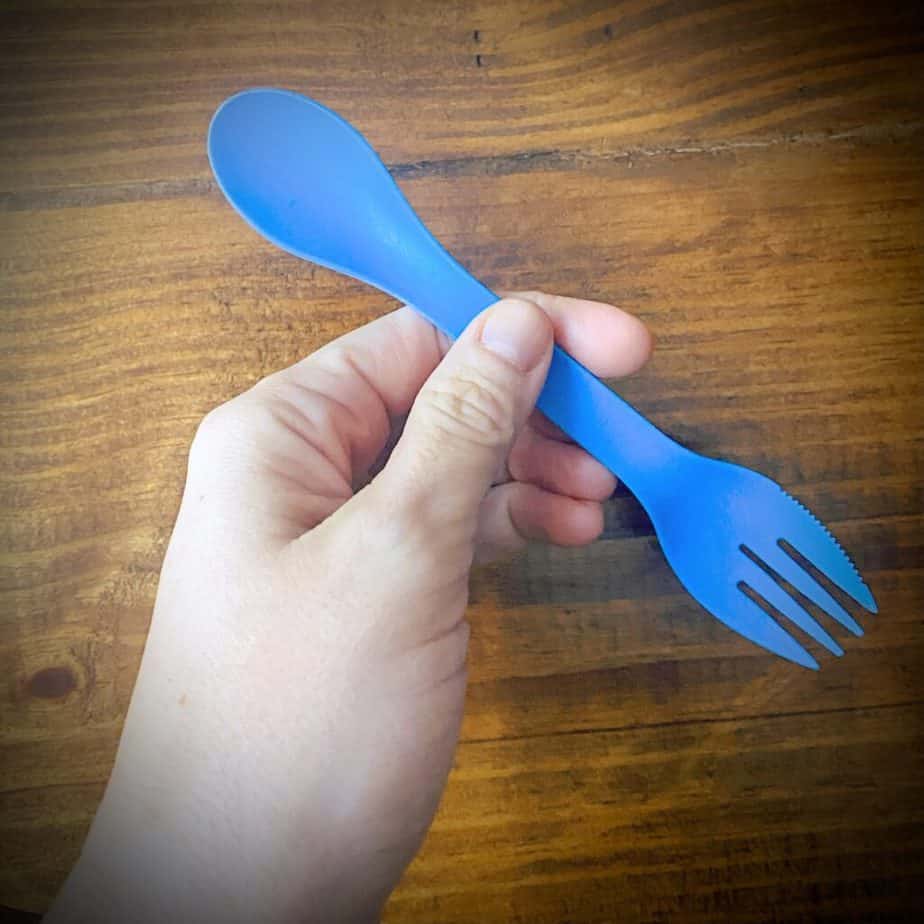 hand holding blue camping utensil with spoon on one side and a serrated fork on the other