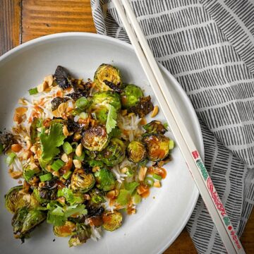 kung pao brussels sprouts in white pasta bowl on brown wood table with a black etched linen and embellished chopsticks.