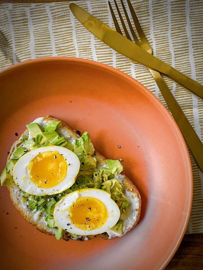 overhead shot of halved turmeric pickled egg on simple avocado toast on a terracotta plate with gold silverware