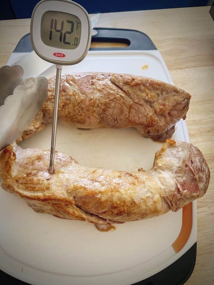 roasted pork tenderloin on a cutting board with a digital meat thermometer showing 142F after pulling from the oven and resting