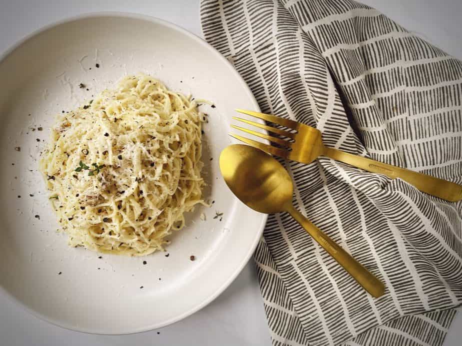 overhead shot of white pasta bowl with a portion of pasta with white clam sauce, gold fork and spoon and a black and white napkin on a white marble countertop