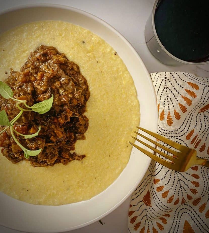 overhead shot of red wine braised short ribs on a bed of parmesan polenta in a white pasta bowl with a glass of red wine and a gold fork
