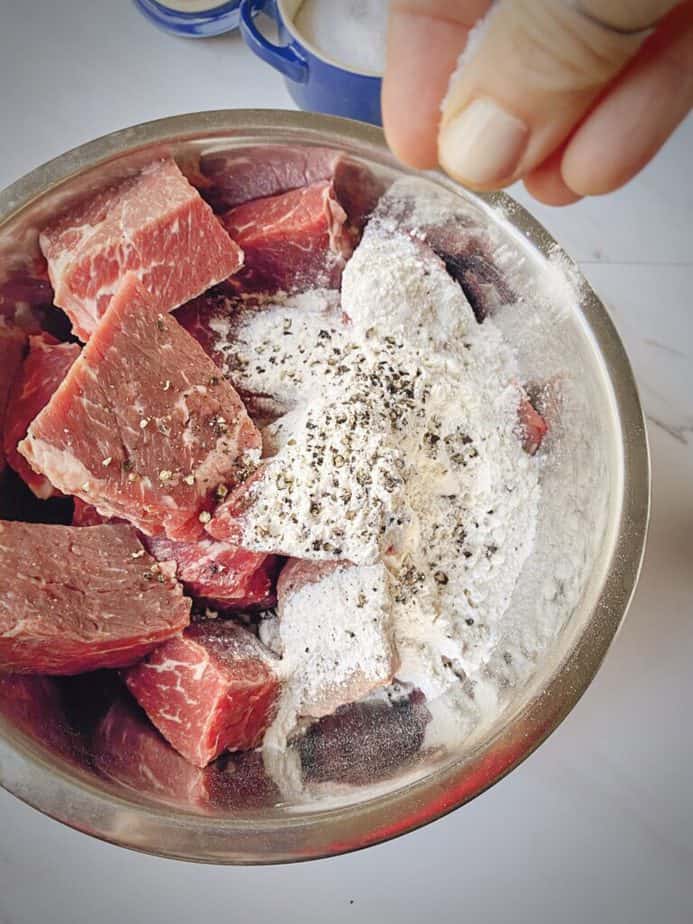 silver bowl with chunks of short ribs, flour, pepper and a hand sprinkling salt