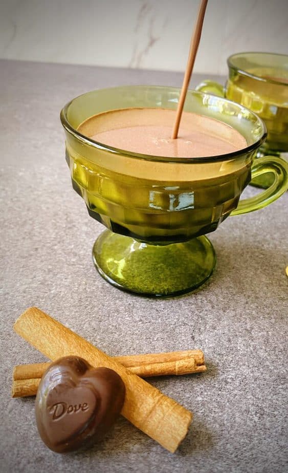 spicy hot chocolate being poured into a green footed mug on a grey table with two cinnamon sticks and a chocolate heart.