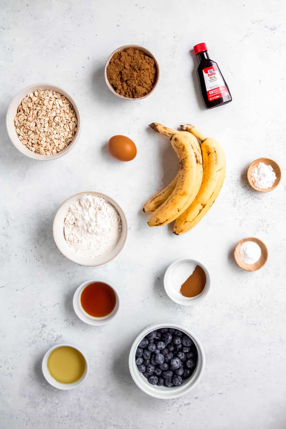 flat lay shot of the ingredients needed to make banana blueberry oatmeal muffins measured out into bowls on a white table.