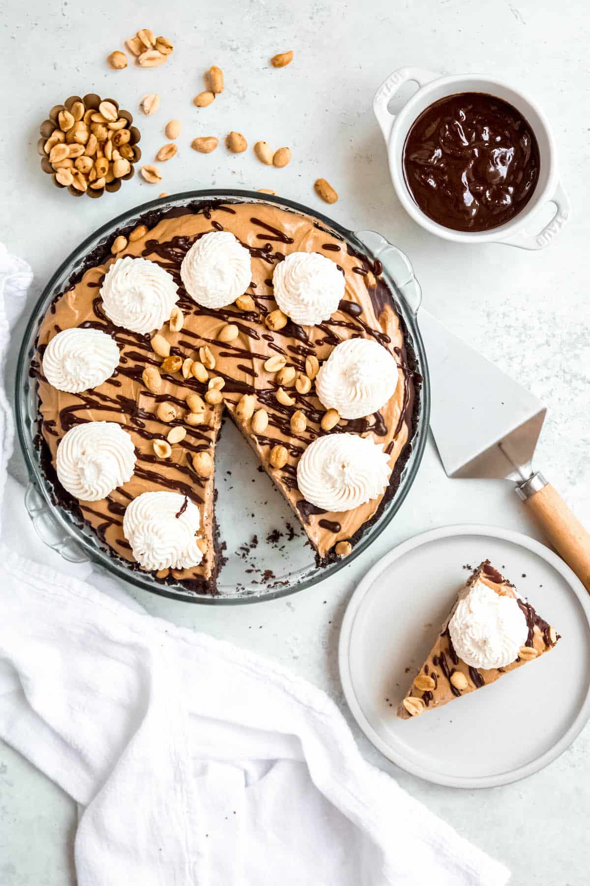 flat lay shot of chocolate peanut butter pie with a slice removed to a white dessert plate plus a bowl of chocolate peanut butter ganache and a ramekin of roasted peanuts on a white table.