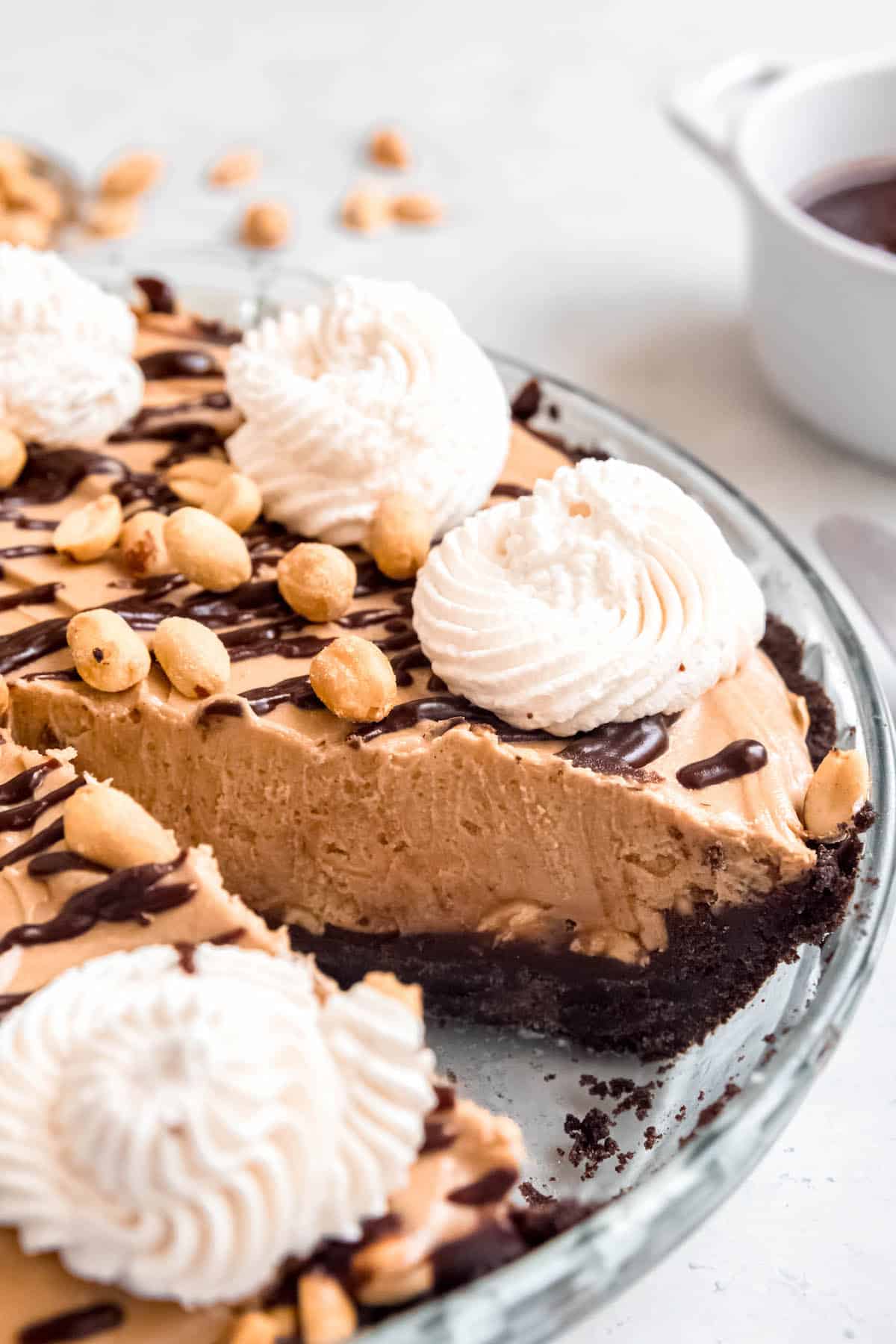 45 degree angle hero shot of the chocolate peanut butter oreo pie with a slice removed to show the layers of oreo crust, chocolate peanut butter ganache, roasted peanuts, peanut butter cream cheese mousse, and all the garnishes.