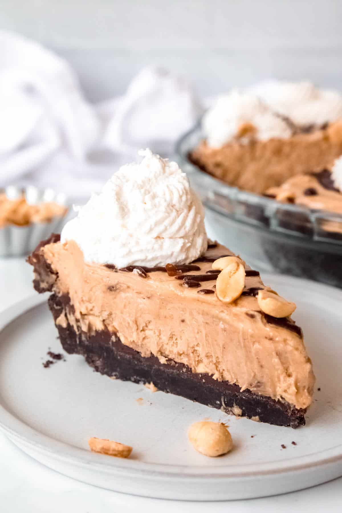 45 degree angle shot of a slice of chocolate peanut butter ganache pie on a white dessert plate with the rest of the pie blurred in the background.