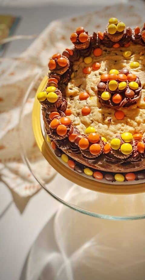 decorated cookie cake with peanut butter chocolate frosting, reeses cups and reeses pieces on a cake stand