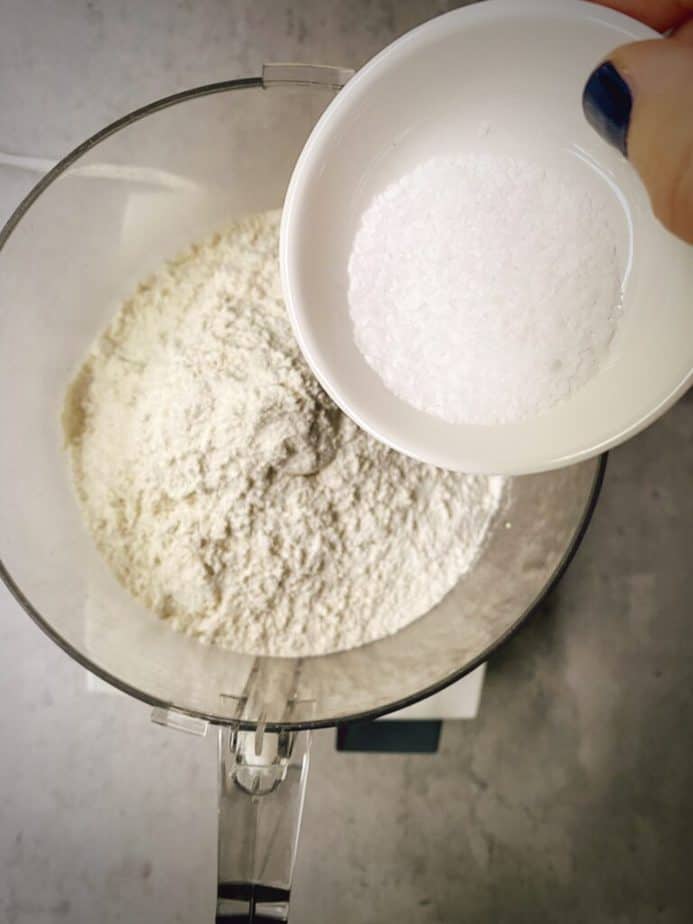 flour in bowl of food processor with hand pouring in the salt