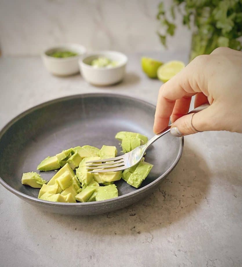 hand using a fork to mash cubes of avocado in a bowl. 