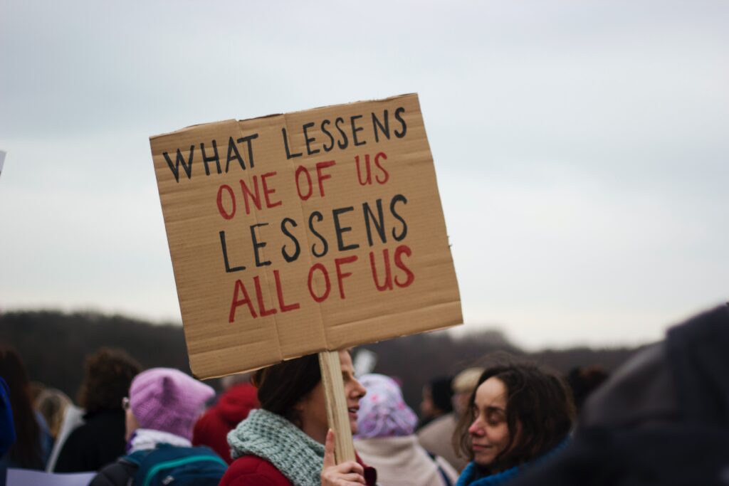 woman holding sign that reads "what lessens one of us lessens all of us."