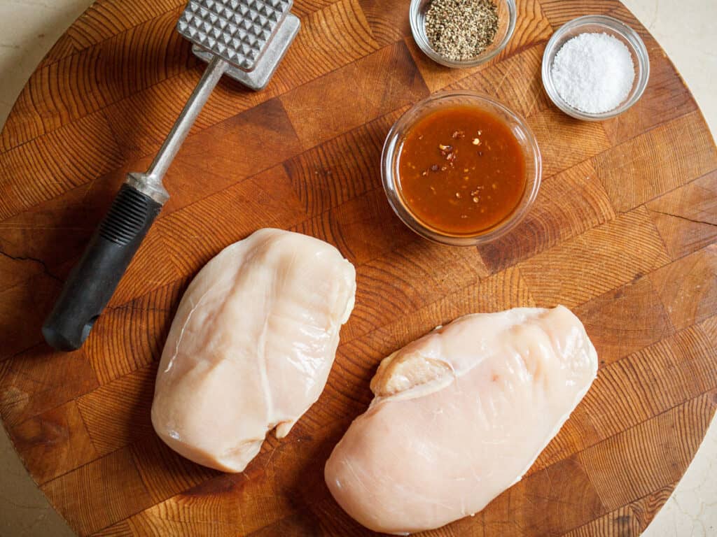 2 chicken breasts on a wooden cutting board with a meat mallet, garlic hot sauce, salt, and pepper.