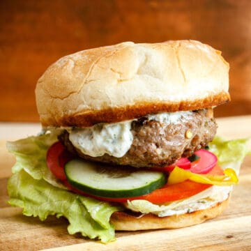 hero image of greek lamb burger on a wooden cutting block with tzatziki dripping down the side.