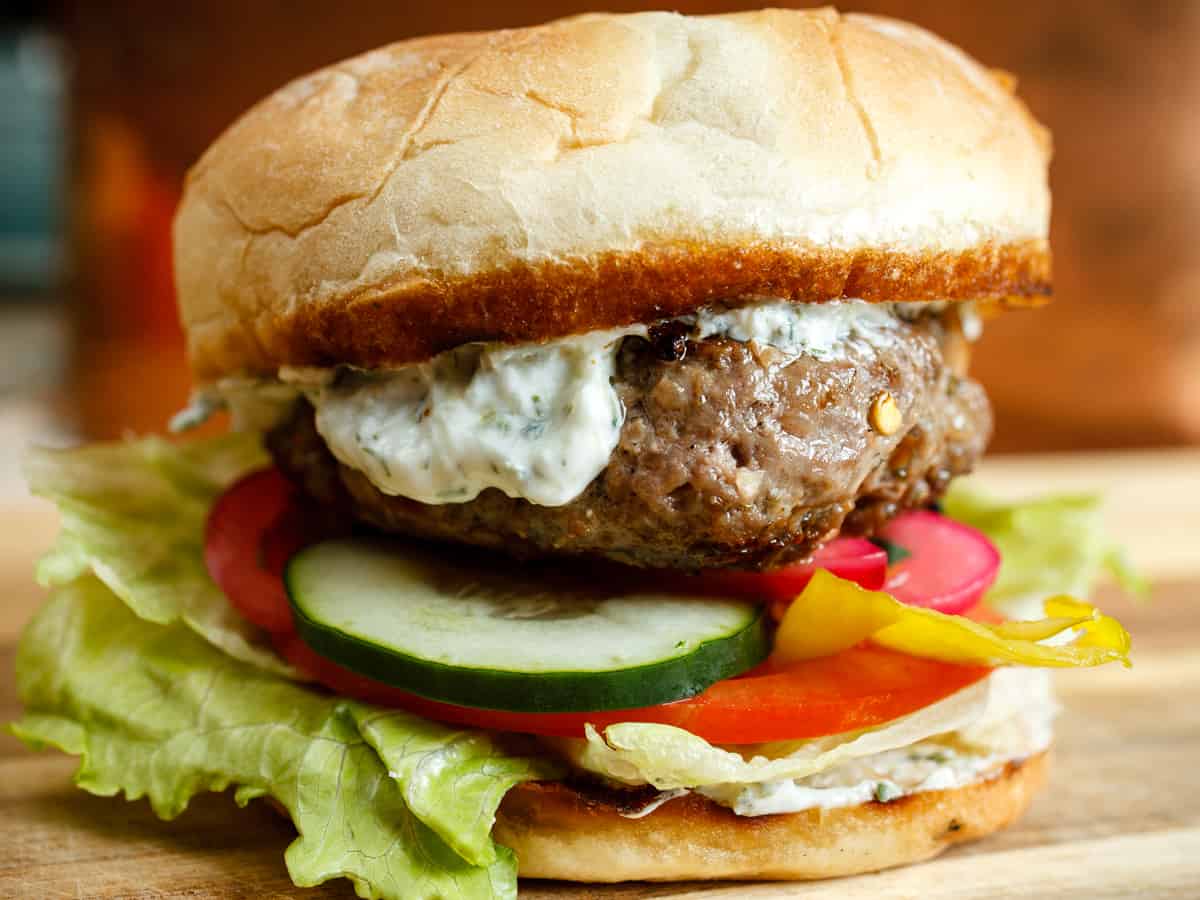 close up shot of an assembled Greek lamb burger with tzatziki, feta, cucumber, pickled red onions, banana peppers, tomato, and lettuce.