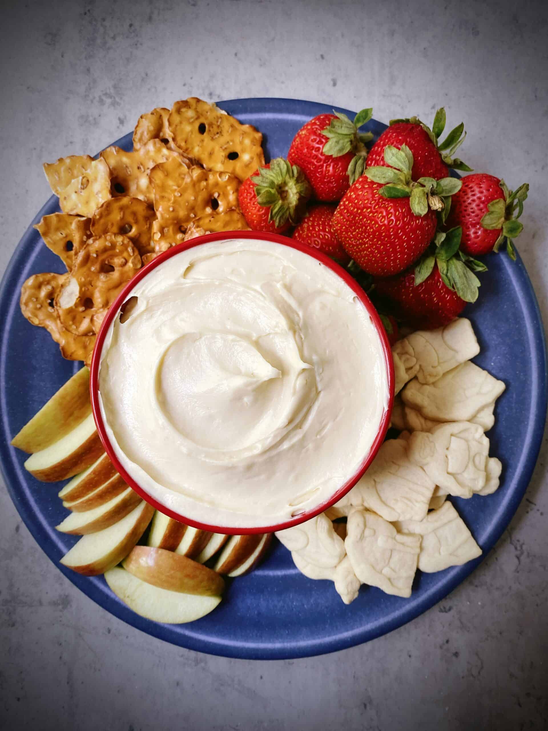 overhead shot of red bowl filled with cream cheese fruit dip on a blue plate with different kinds of fruit and crackers.