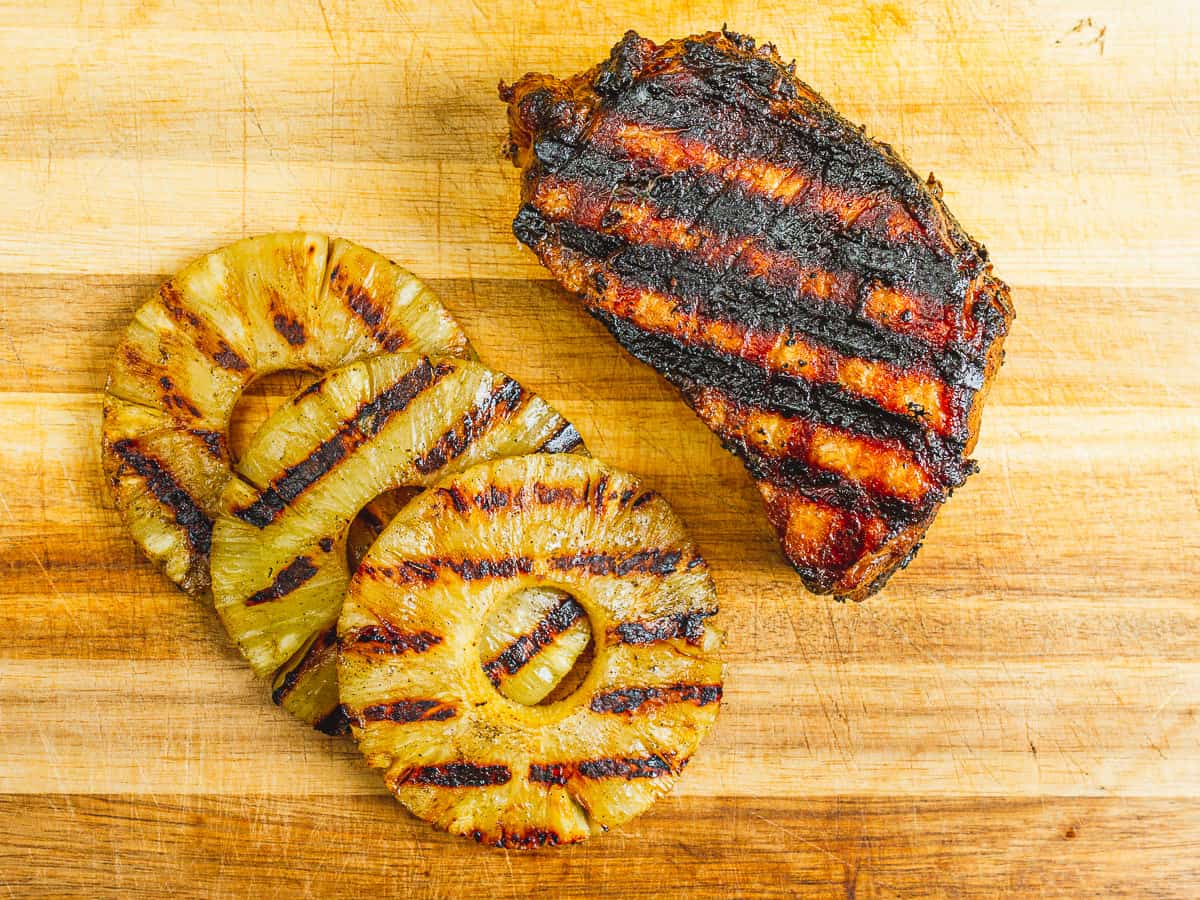 three rings of grilled pineapple and a grilled hawaiian pork chop on the cutting board.