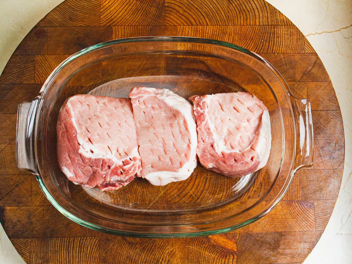 three thick cut pork chops in a glass dish after being pricked with a knife all over.