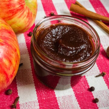 no peel apple butter in a small mason jar on a red and white checked cloth with whole apples and cinnamon sticks in the background.