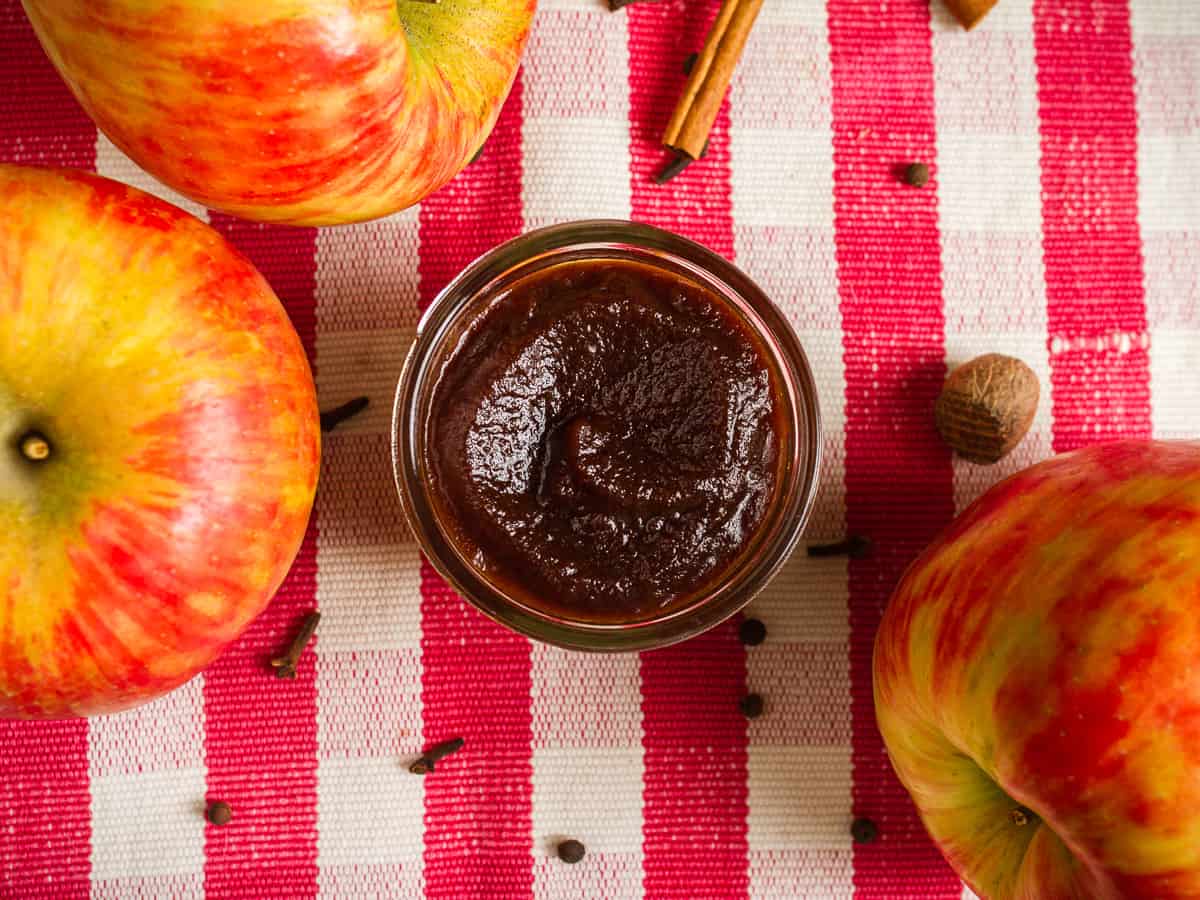 hero shot of slow cooker apple butter in a mason jar with whole apples, whole clove, and a whole nutmeg scattered around.