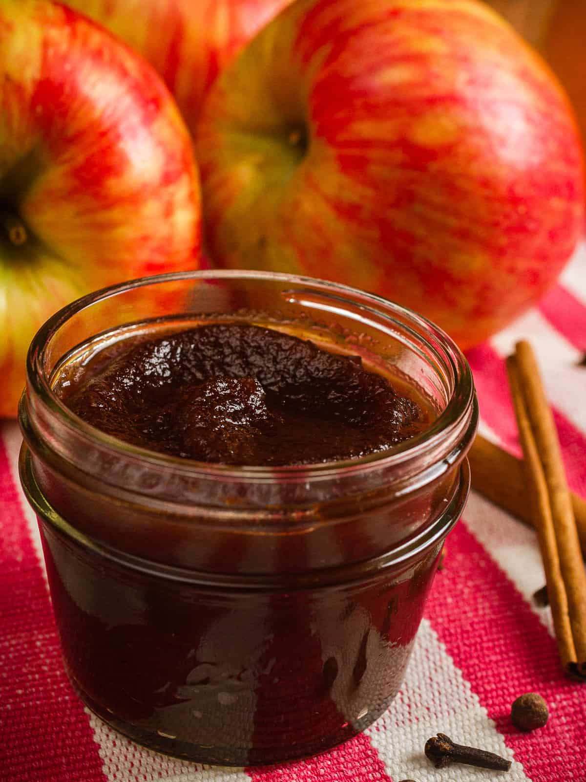 vertical shot of no peel apple butter in a small mason jar on a red and white checked cloth with whole apples and cinnamon sticks in the background.