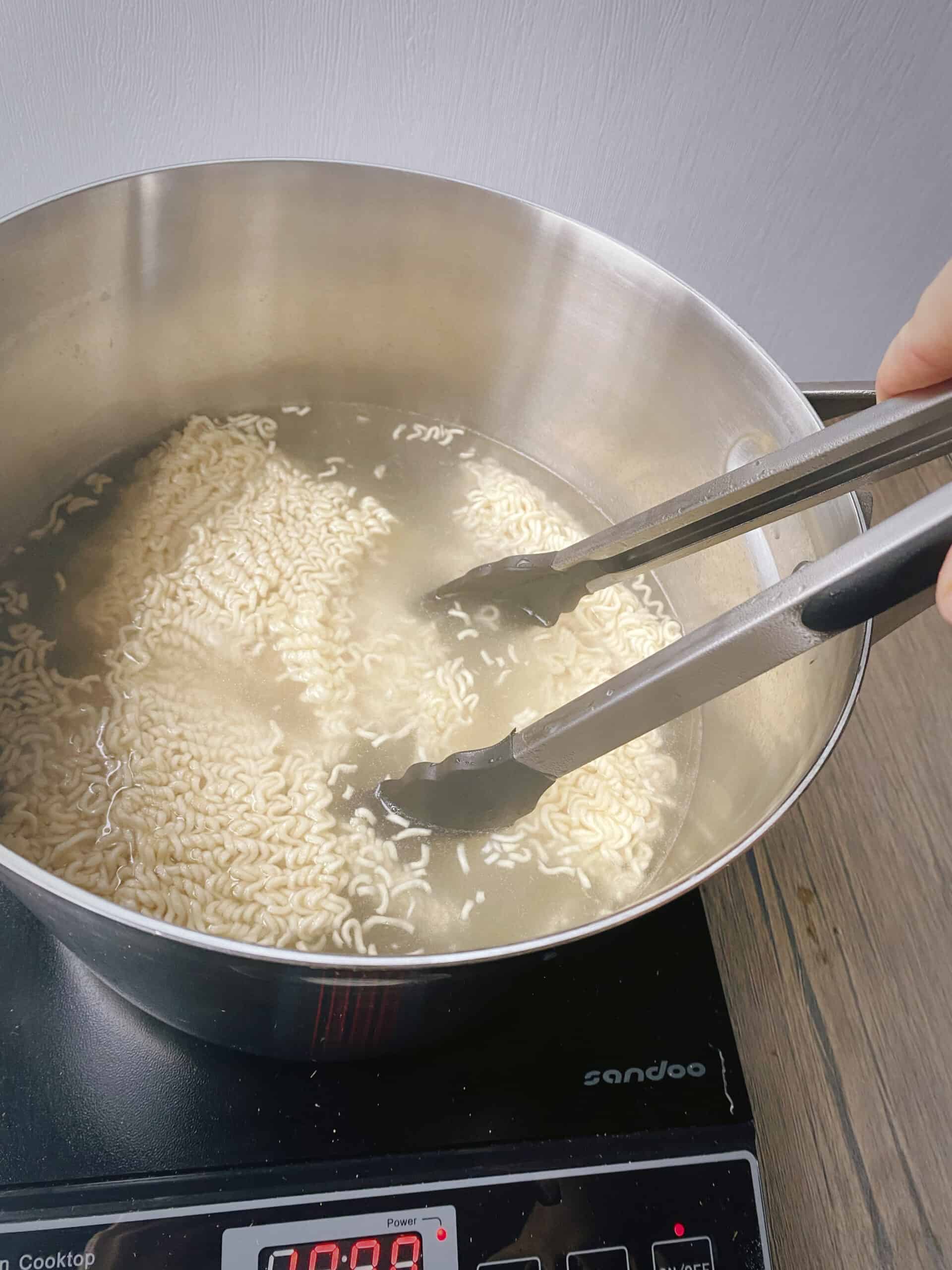 tongs stirring the ramen noodles in the boiling water as they cook.