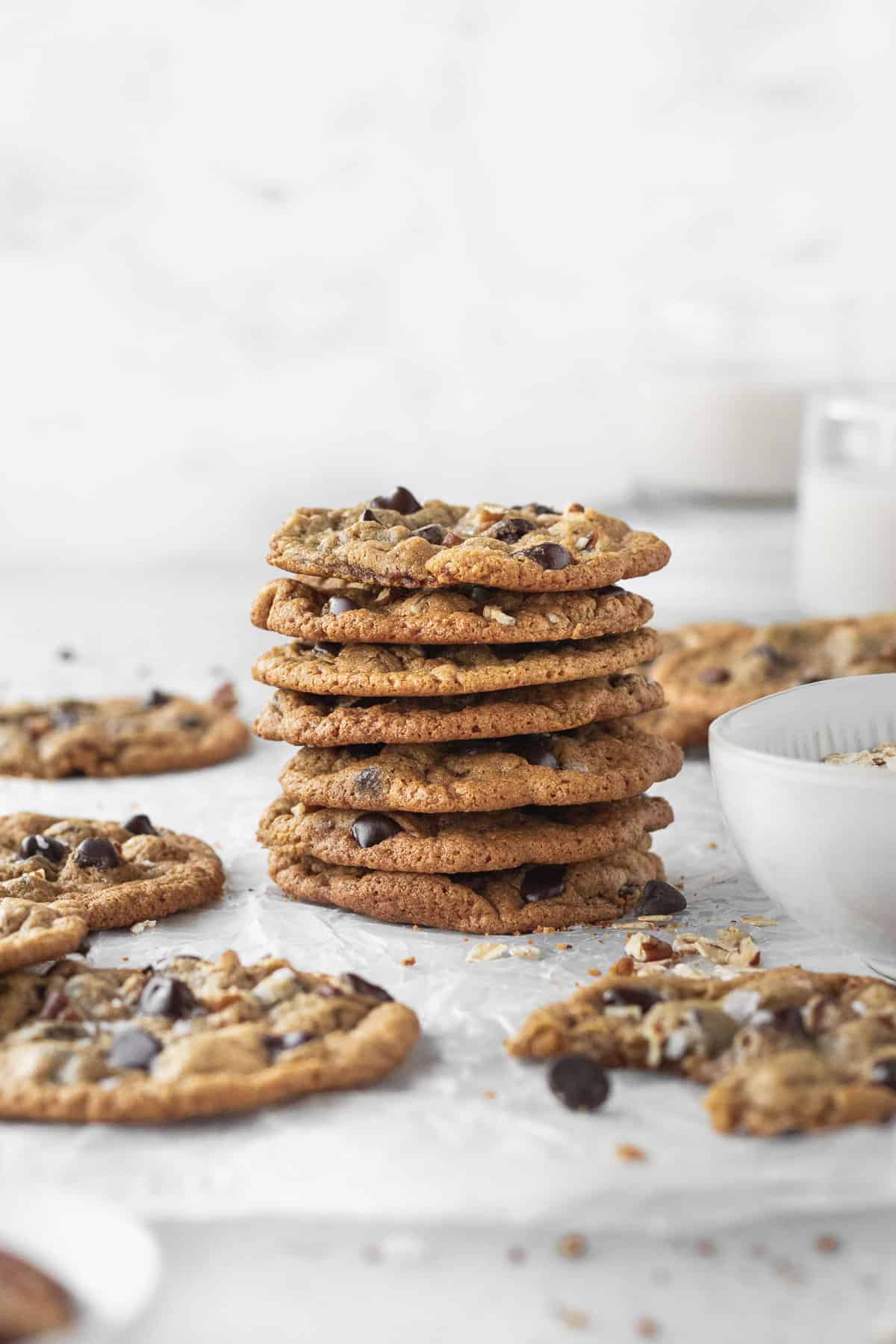 sidesways shot of a stack of thin and crispy oat flour chocolate chip cookies with a glass of milk in the background.