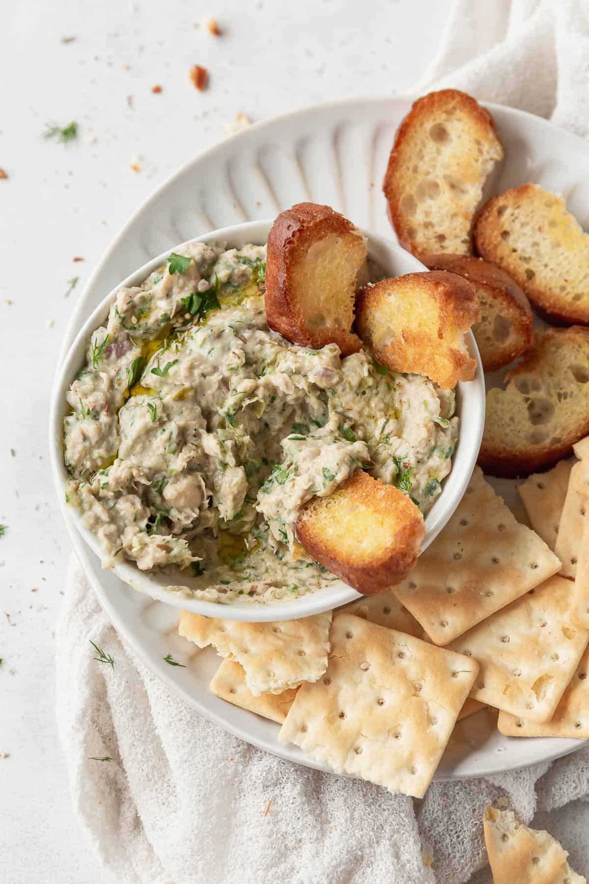 pieces of crostini tucked into a bowl of mediterranean tuna dip with white beans.