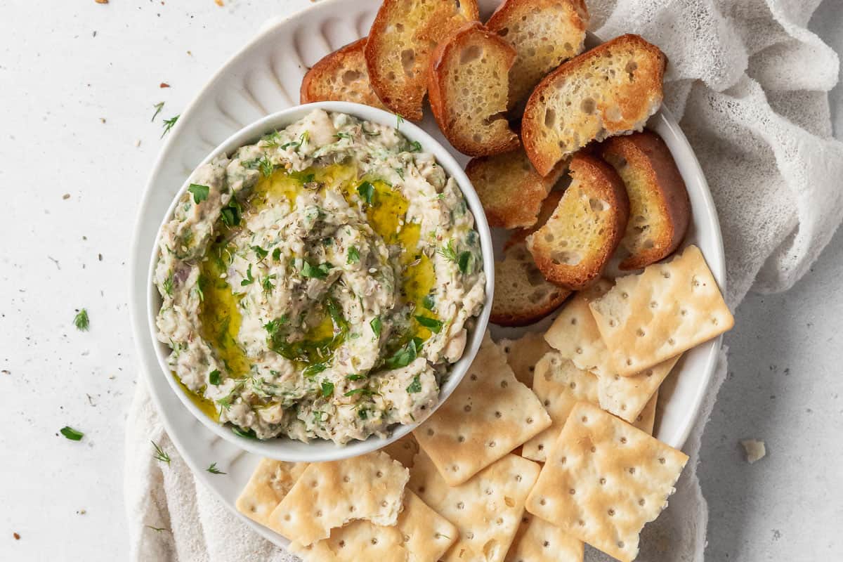 horizontal overhead hero of a serving platter filled with crostini and saltines with a bowl of white bean tuna dip.