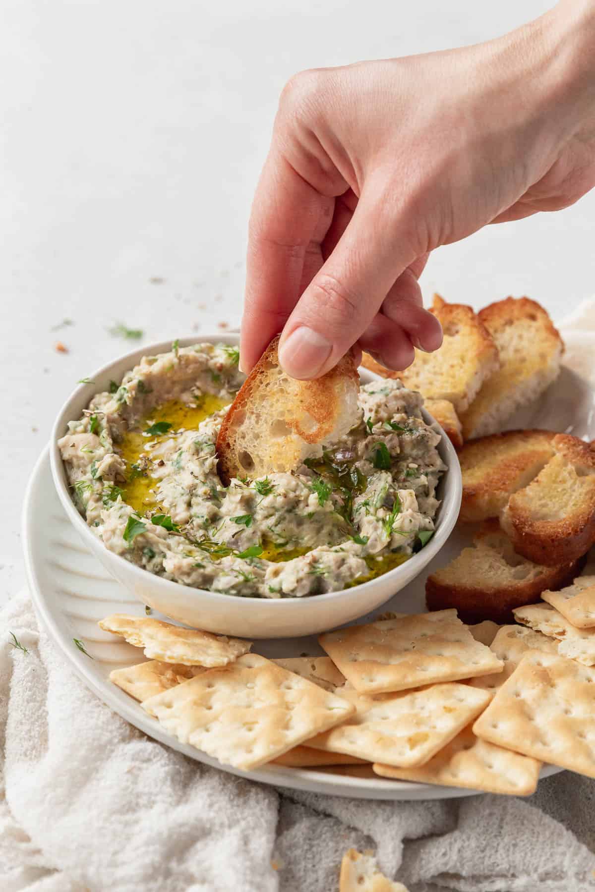 vertical hero of a hand dipping a crostini into a bowl of white bean tuna dip with no cream cheese.