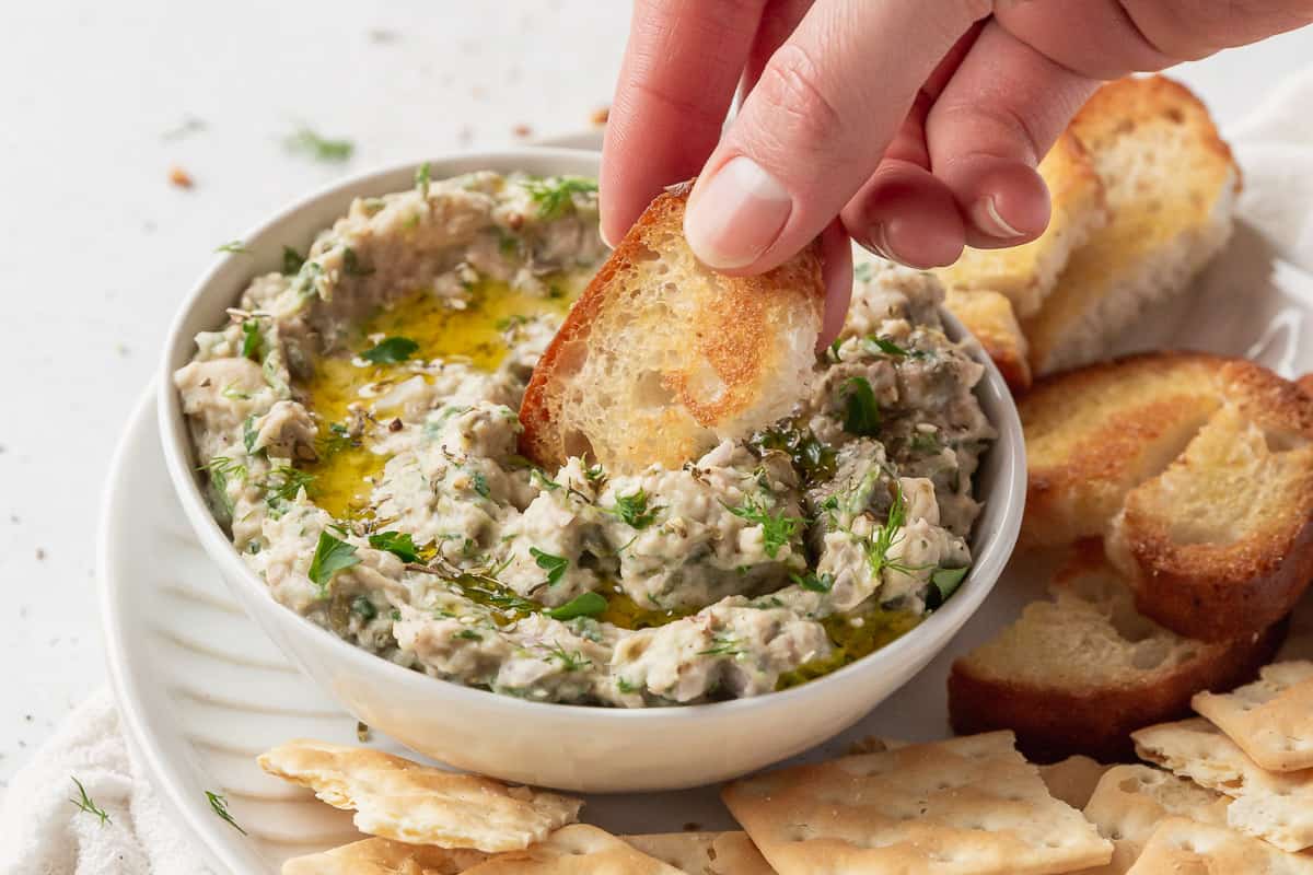 closeup of a hand dipping a toasted crostini into healthy gluten-free tuna dip with white beans.